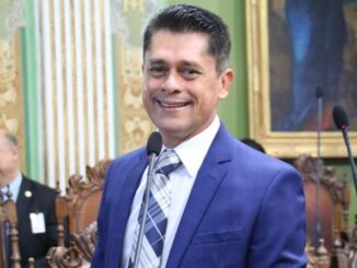 Vereador Luiz Carlos sorri durante sessão na Câmara Municipal de Salvador, usando terno azul e gravata listrada.