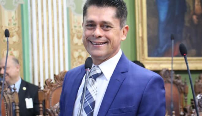 Vereador Luiz Carlos destaca conquistas do Republicanos em Salvador Vereador Luiz Carlos sorri durante sessão na Câmara Municipal de Salvador, usando terno azul e gravata listrada.