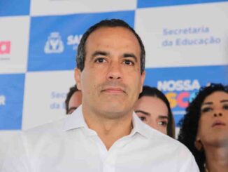 Prefeito de Salvador em evento institucional da prefeitura, diante de painel com logotipos da gestão municipal.