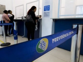 Fila de atendimento em agência do INSS, com faixa azul identificada como “Previdência Social” em destaque no primeiro plano.