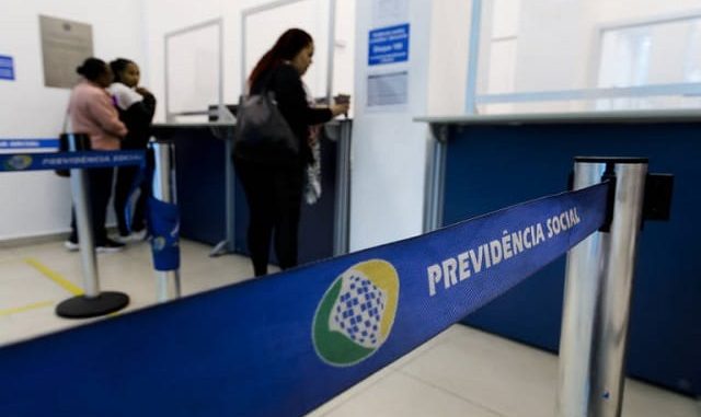 Atendimento em agência da Previdência Social no Brasil Fila de atendimento em agência do INSS, com faixa azul identificada como “Previdência Social” em destaque no primeiro plano.