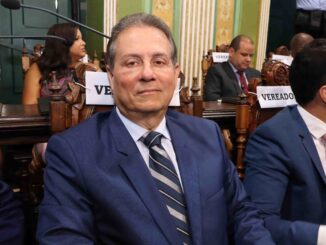 Vereador sentado em plenário da Câmara Municipal de Salvador, usando terno azul e gravata listrada.