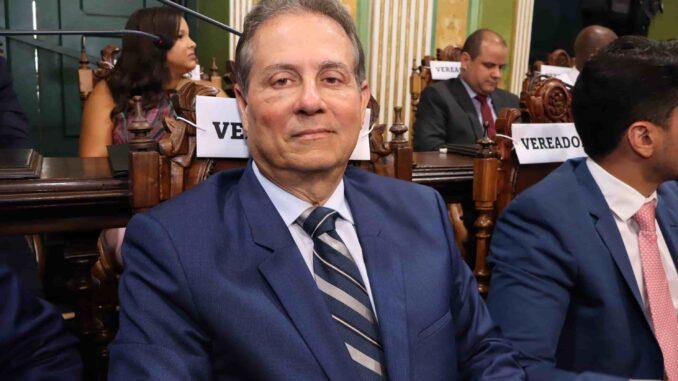 Vereador sentado em plenário da Câmara Municipal de Salvador, usando terno azul e gravata listrada.