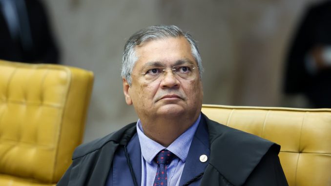 Ministro Flávio Dino sentado em sua cadeira no plenário do STF, usando toga preta, camisa azul e gravata vermelha com pontos azuis.