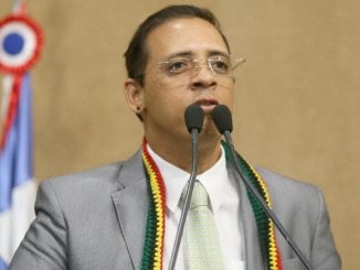 Homem de terno cinza e gravata clara fala ao microfone duplo em plenário, com bandeira ao fundo.