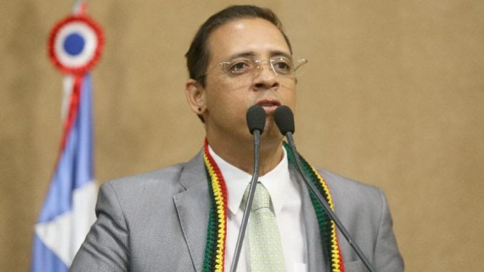 Homem de terno cinza e gravata clara fala ao microfone duplo em plenário, com bandeira ao fundo.