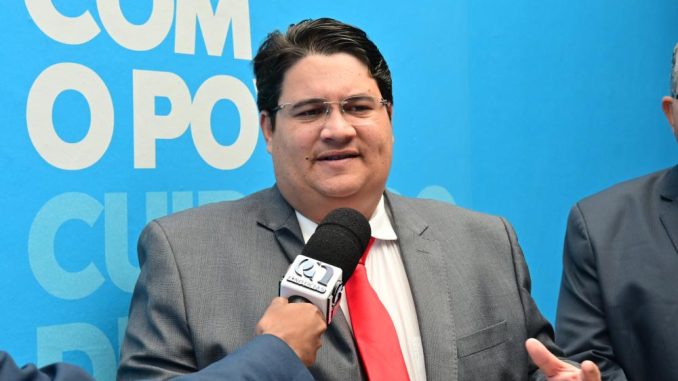 Vereador Tagner Cerqueira (PT) fala à imprensa, usando terno cinza e gravata vermelha, durante entrevista com microfone em frente a painel azul.