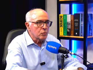 Homem de óculos e camisa azul fala ao microfone durante entrevista em estúdio, com livros ao fundo em prateleira iluminada.