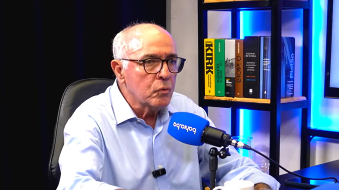 Homem de óculos e camisa azul fala ao microfone durante entrevista em estúdio, com livros ao fundo em prateleira iluminada.