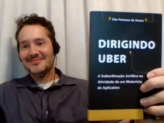 Homem segurando o livro “Dirigindo Uber: A Subordinação Jurídica na Atividade de um Motorista de Aplicativo”, com headset e sorriso, em frente a uma cortina branca.