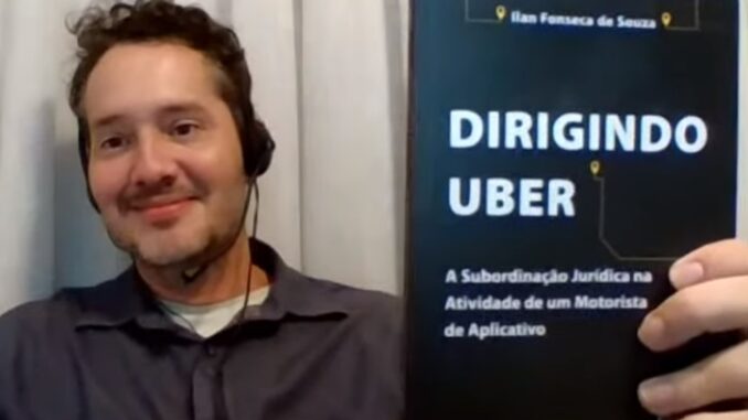 Procurador do Trabalho Ilan Fonseca apresenta livro sobre motoristas de aplicativos Homem segurando o livro “Dirigindo Uber: A Subordinação Jurídica na Atividade de um Motorista de Aplicativo”, com headset e sorriso, em frente a uma cortina branca.