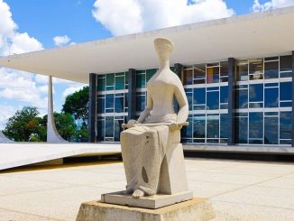 Estátua da Justiça em frente ao prédio do Supremo Tribunal Federal em Brasília, sob céu azul com nuvens