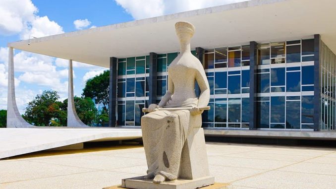 Estátua da Justiça em frente ao prédio do Supremo Tribunal Federal em Brasília, sob céu azul com nuvens