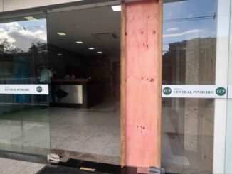 Fachada de prédio com vidro quebrado e parte coberta por madeira após disparo de bala perdida.