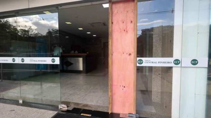 Fachada de prédio com vidro quebrado e parte coberta por madeira após disparo de bala perdida.